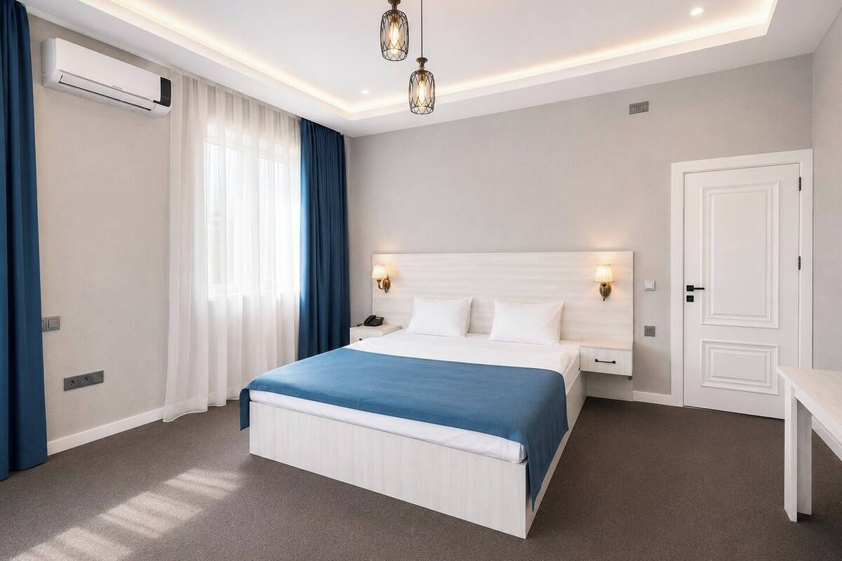 A hotel room with a bed, bedside lamps, a desk, and an air conditioning unit.