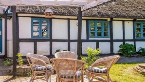 A traditional half-timbered house with a thatched roof, a covered patio with wicker chairs, and a small garden.