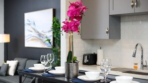 A modern dining area with a table set for two, a vase of pink flowers, and a kitchen in the background. - Spacious Modern 1-Bedroom, Near Riverwalk & Dome (San Antonio)