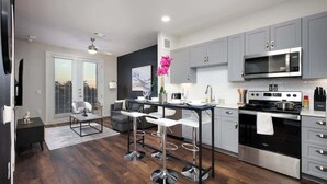 A modern kitchen with grey cabinets, a stainless steel stove, and a black microwave. There are three white bar stools and a dining table with a vase of pink flowers. - Spacious Modern 1-Bedroom, Near Riverwalk & Dome (San Antonio)