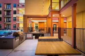 An outdoor seating area with a wooden table and grill, surrounded by a pool and a building with balconies. - Modern 2-Bedroom Getaway Near Riverwalk & Dome (San Antonio)
