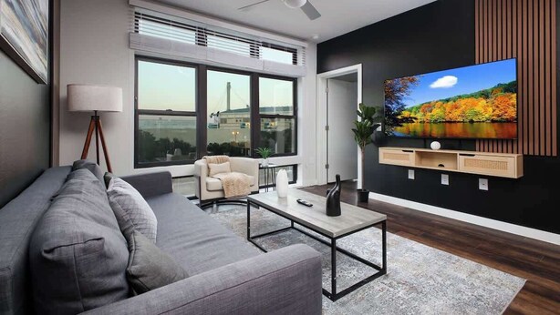 Una sala de estar moderna con un sofá gris, una mesa de centro, un televisor montado en la pared y un ventanal con vistas a un puente.