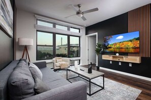 Una sala de estar moderna con un sofá gris, una mesa de centro, un televisor montado en la pared y un ventanal con vistas a un puente.