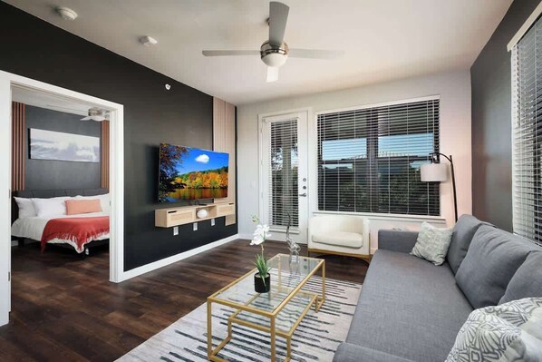 A modern living room with a grey sofa, a coffee table, a TV, and a large window with blinds.