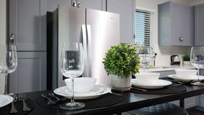 A modern kitchen with grey cabinets, a stainless steel refrigerator, and a dining table set for two with wine glasses and plates.