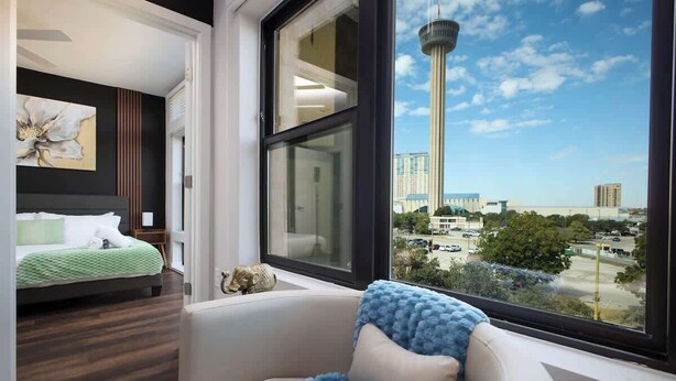 A modern hotel room with a large window, a bed, a sofa, and a view of a tower and cityscape. - Downtown 1-Bedroom Stay with Gym and Free Parking (San Antonio)
