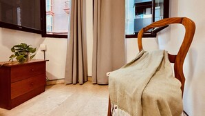 A room with a wooden chair covered with a beige blanket, a wooden dresser with a potted plant, and a window with curtains. - Click&Guest - 1Naval Nautical Ap. in Gran Canaria (Las Palmas de Gran Canaria)