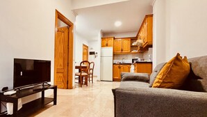 A living room with a grey sofa, a television on a stand, and a small table. There is a kitchen in the background with wooden cabinets and a refrigerator. - Click&Guest - 1Naval Nautical Ap. in Gran Canaria (Las Palmas de Gran Canaria)
