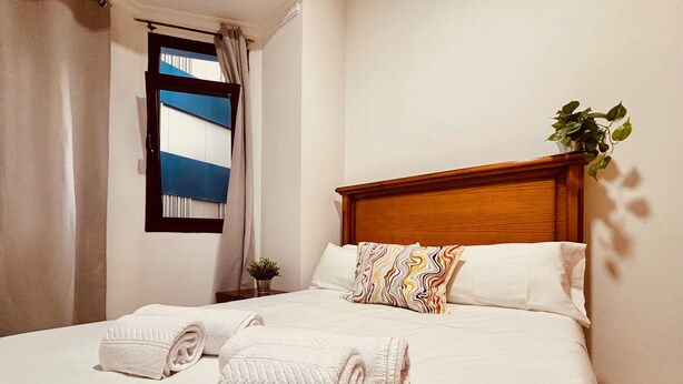 A neatly made bed with white linens and two rolled towels, a wooden headboard, a small bedside table with a plant, and a window with blue curtains. - Click&Guest - 1Naval Nautical Ap. in Gran Canaria (Las Palmas de Gran Canaria)