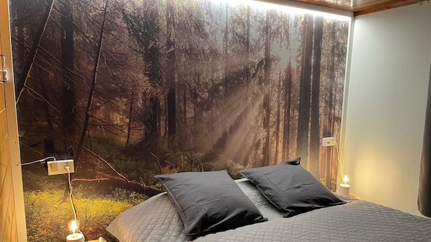 A bedroom with a large forest mural on the wall, a bed with a quilted cover, two pillows, a bedside table with a lamp, and a power outlet. - Relax and take your time at Chalet du temps suspendu (Rendeux)