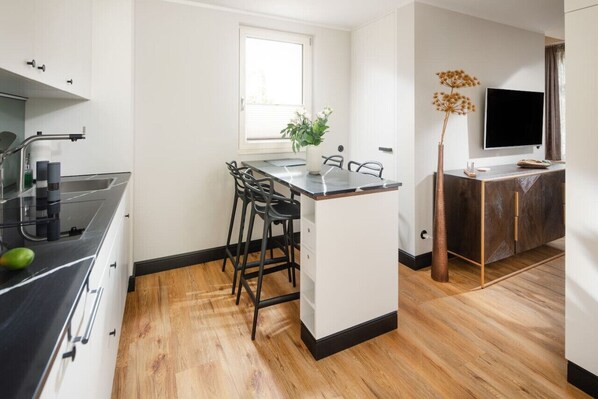 A modern kitchen with a black countertop, white cabinets, and a wooden floor. There is a window above the sink, a vase with flowers on the island, and a flat-screen TV mounted on the wall.