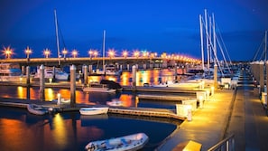 A marina with boats docked, a bridge overhead, and a well-lit waterfront.