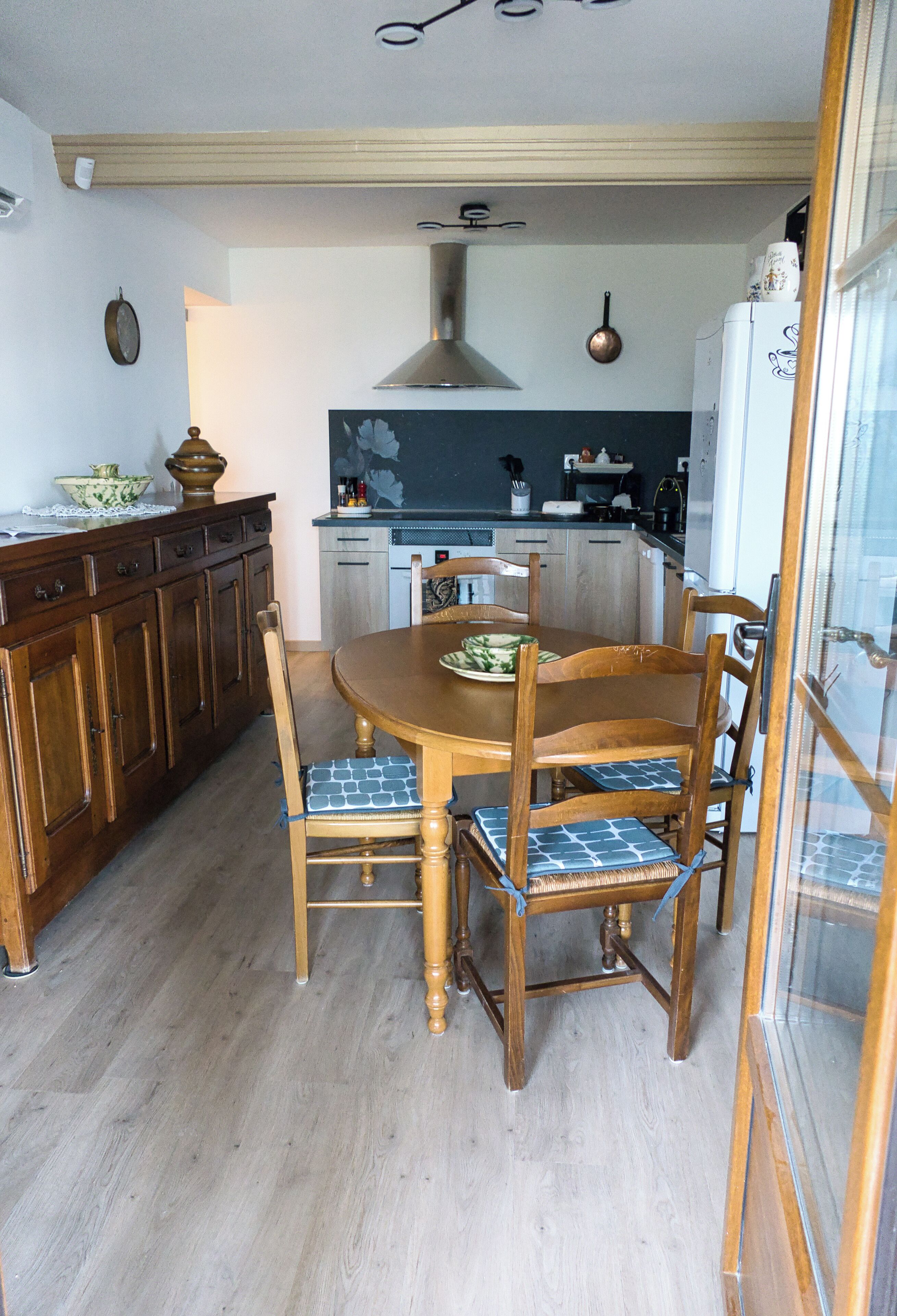 A dining area with a wooden table and chairs, a kitchen in the background, and a wooden floor.