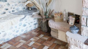 A corner of a room with a stone wall, a brick floor, a bench, a vase with dried plants, wall sconces, and a candle holder.