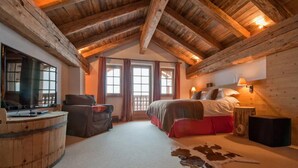 A cozy room with a wooden ceiling, a bed with a red bedspread, a leather chair, a wooden barrel, and a large window with curtains. - Chalet Baker Vacation Home (Pettneu am Arlberg)