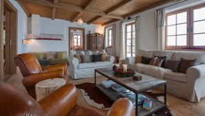 A cozy living room with wooden beams, a leather armchair, a sofa, and a coffee table with books and flowers. - Chalet Baker Vacation Home (Pettneu am Arlberg)