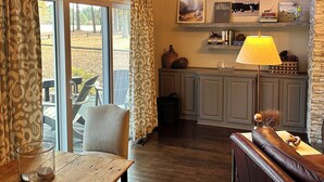 Una sala de estar acogedora con una mesa de madera, un sofá de cuero y una mesa de comedor. Hay una zona de cocina con una lámpara y un estante con libros y objetos decorativos. Amplias ventanas ofrecen vistas al exterior.