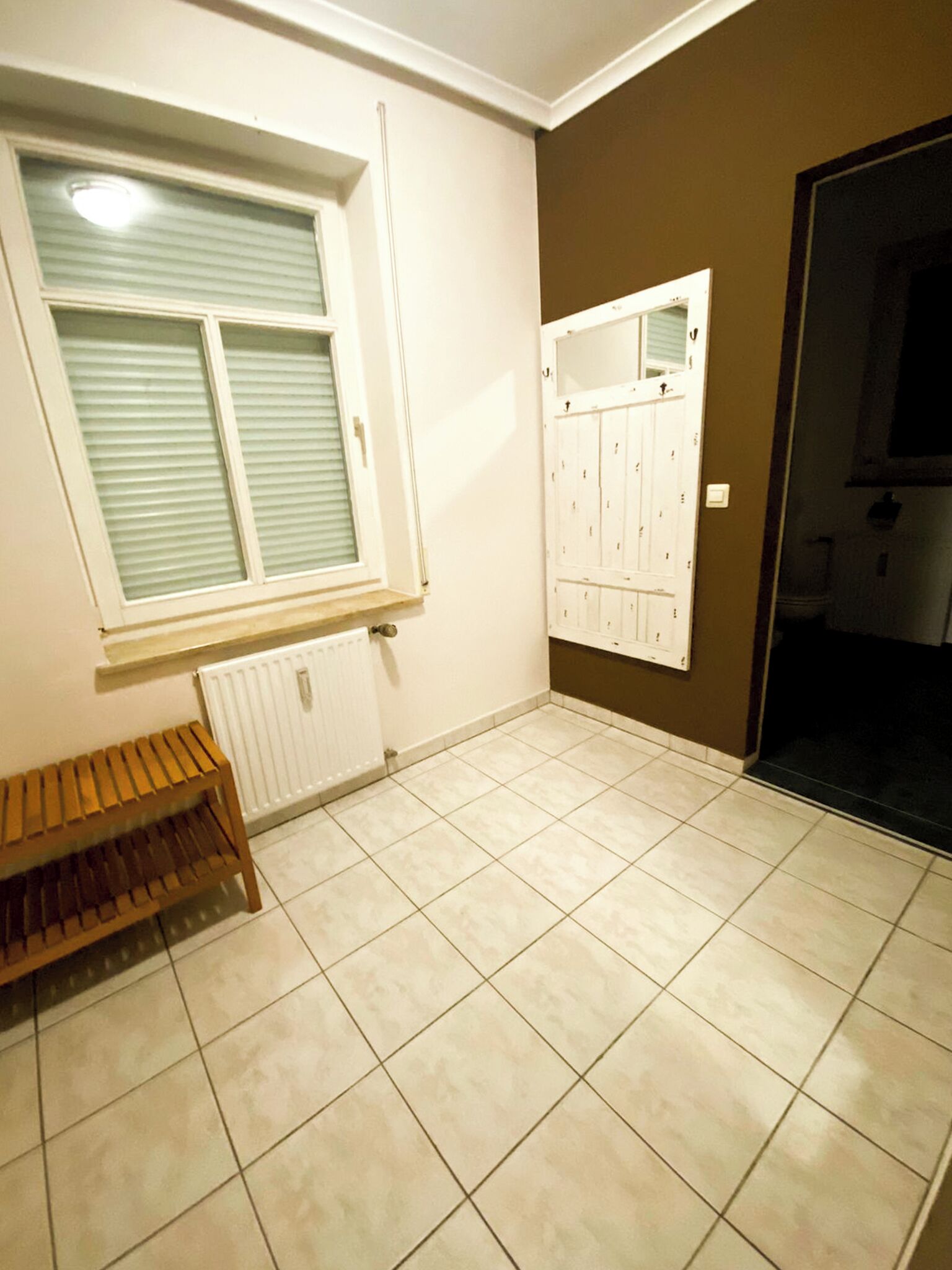 Una habitación con un radiador, un banco, una ventana con persianas y una puerta que da a una zona oscura.