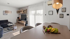 A modern living room with a sofa, coffee table, and dining table with fruit. There is a TV, shelves, and a large window with curtains. - Premium Apartment Seaview (Porec)