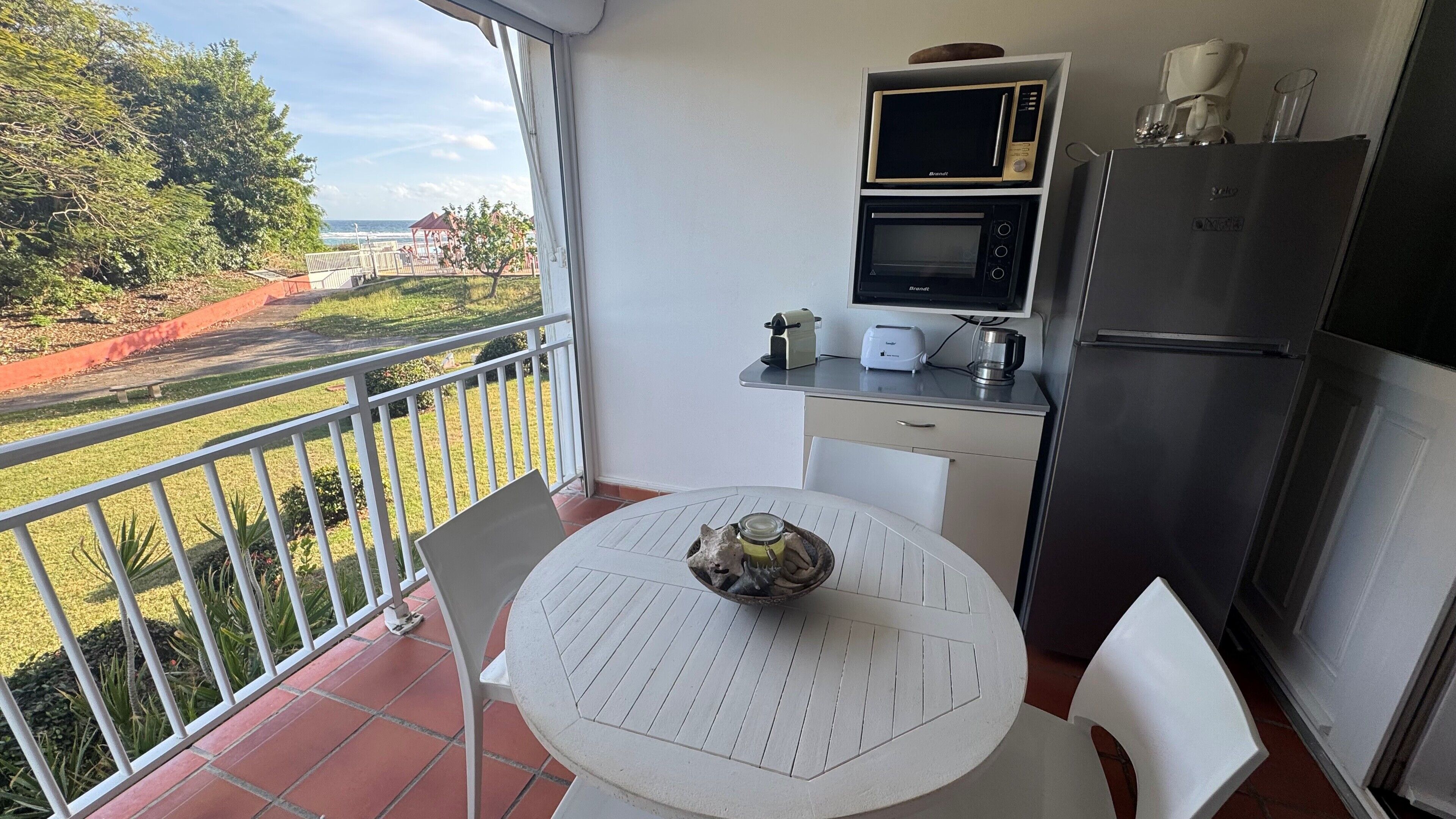 A balcony with a table and chairs, a small kitchen area with a microwave, toaster, and refrigerator, and a view of a landscaped garden and distant buildings.