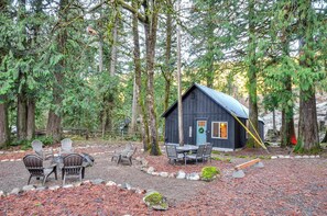 A rustic cabin with a wooden exterior, surrounded by a forest. There is a table and chairs set outside, and a swing in the yard.