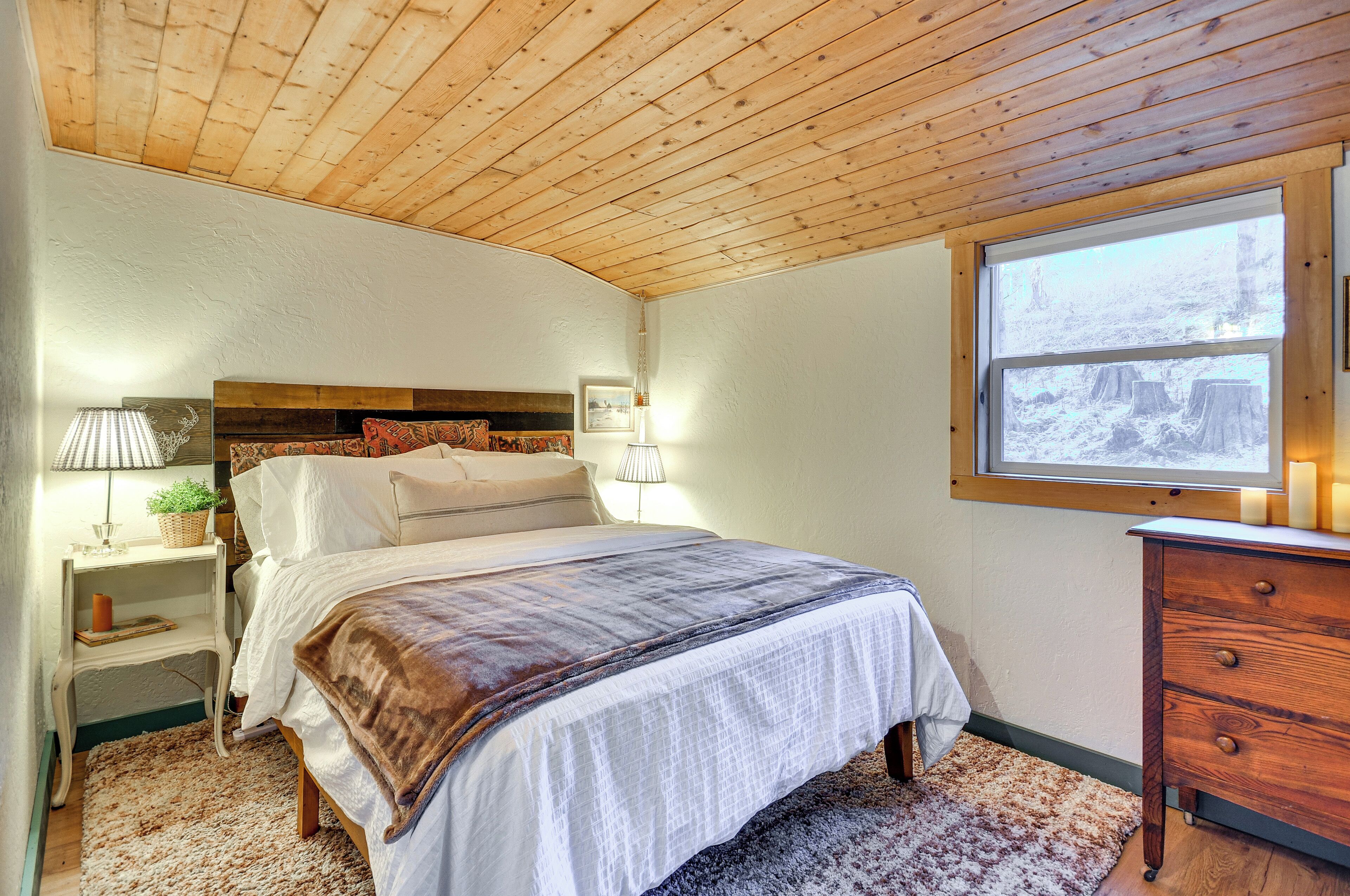 A bedroom with a wooden ceiling, a bed with a white and brown blanket, two bedside lamps, a nightstand with a lamp and a plant, and a dresser.