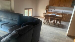 Un salon avec un canapé d’angle gris, un coin repas avec des chaises en bois et une cuisine avec des meubles en bois et un plan de travail.