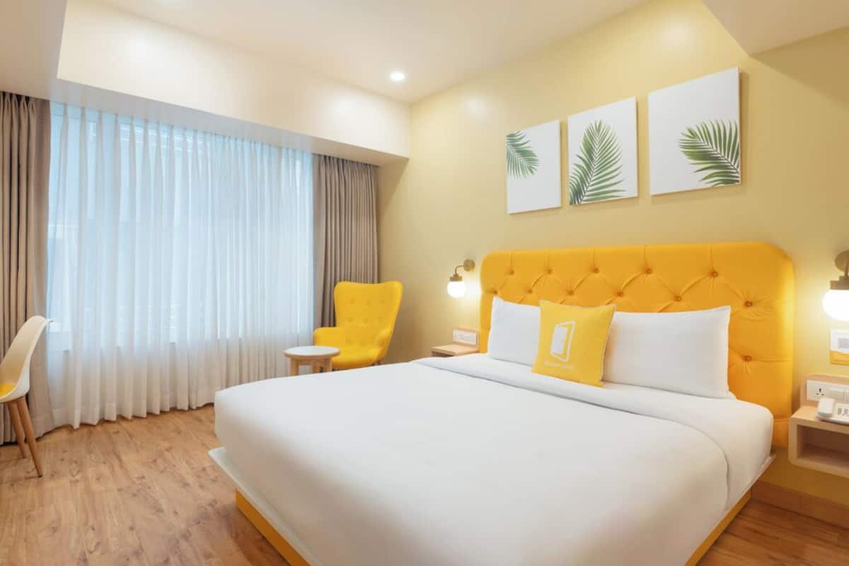 A hotel room with a large bed, a yellow headboard, and a window with curtains. There are two wall-mounted paintings and a small table with a chair.