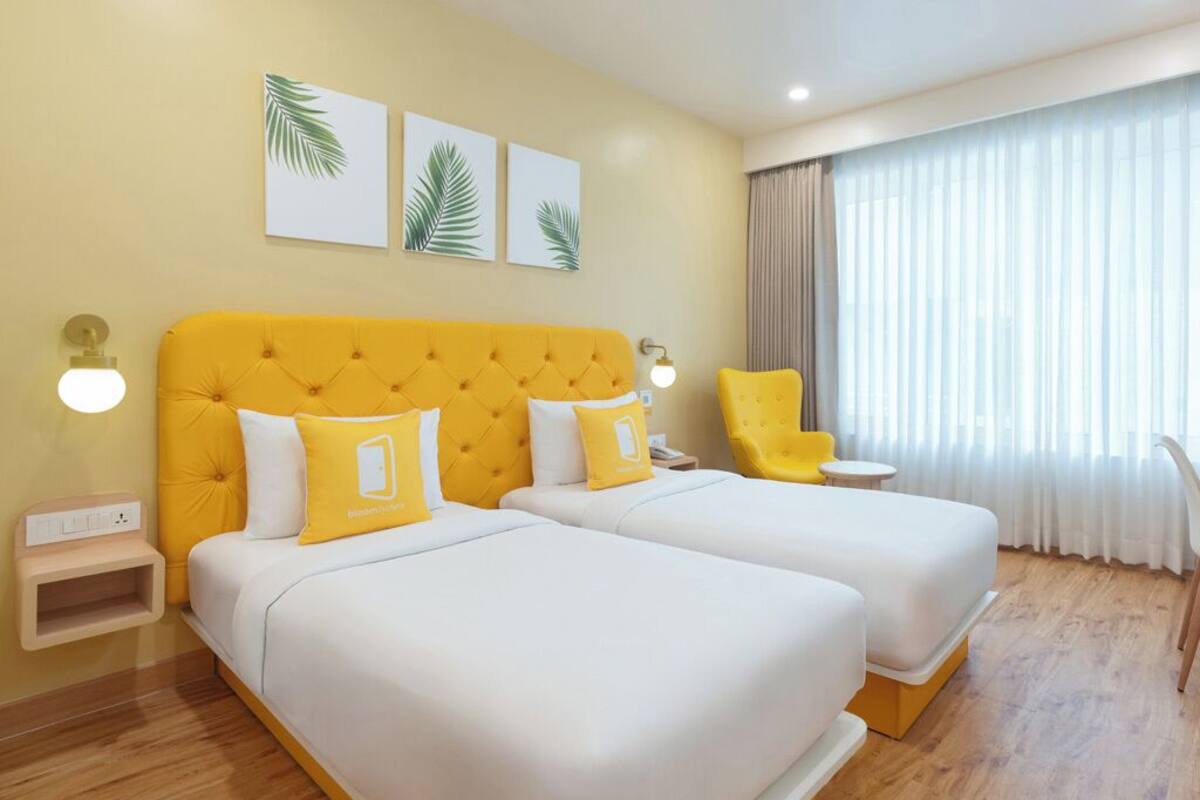 A hotel room with two beds, a yellow headboard, white bedding, a small table, a chair, and a window with curtains.