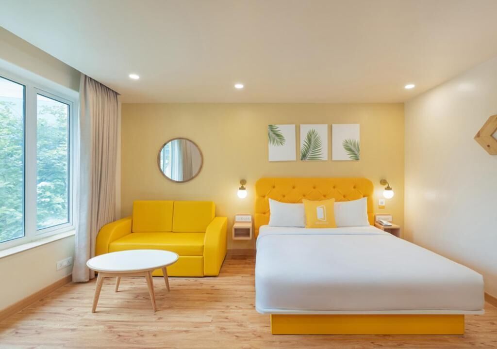 Une chambre d’hôtel avec un lit, un canapé jaune, un miroir rond et une petite table.