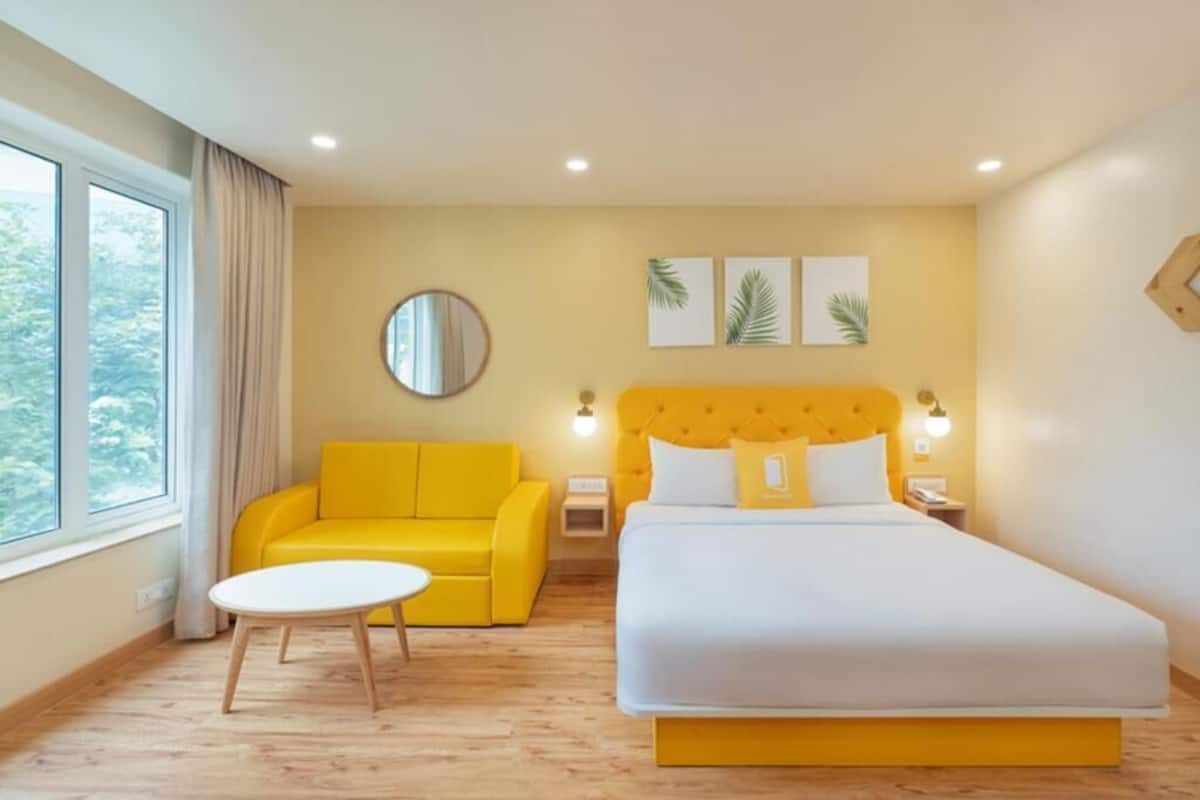 A hotel room with a bed, a yellow sofa, a round mirror, and a small table.