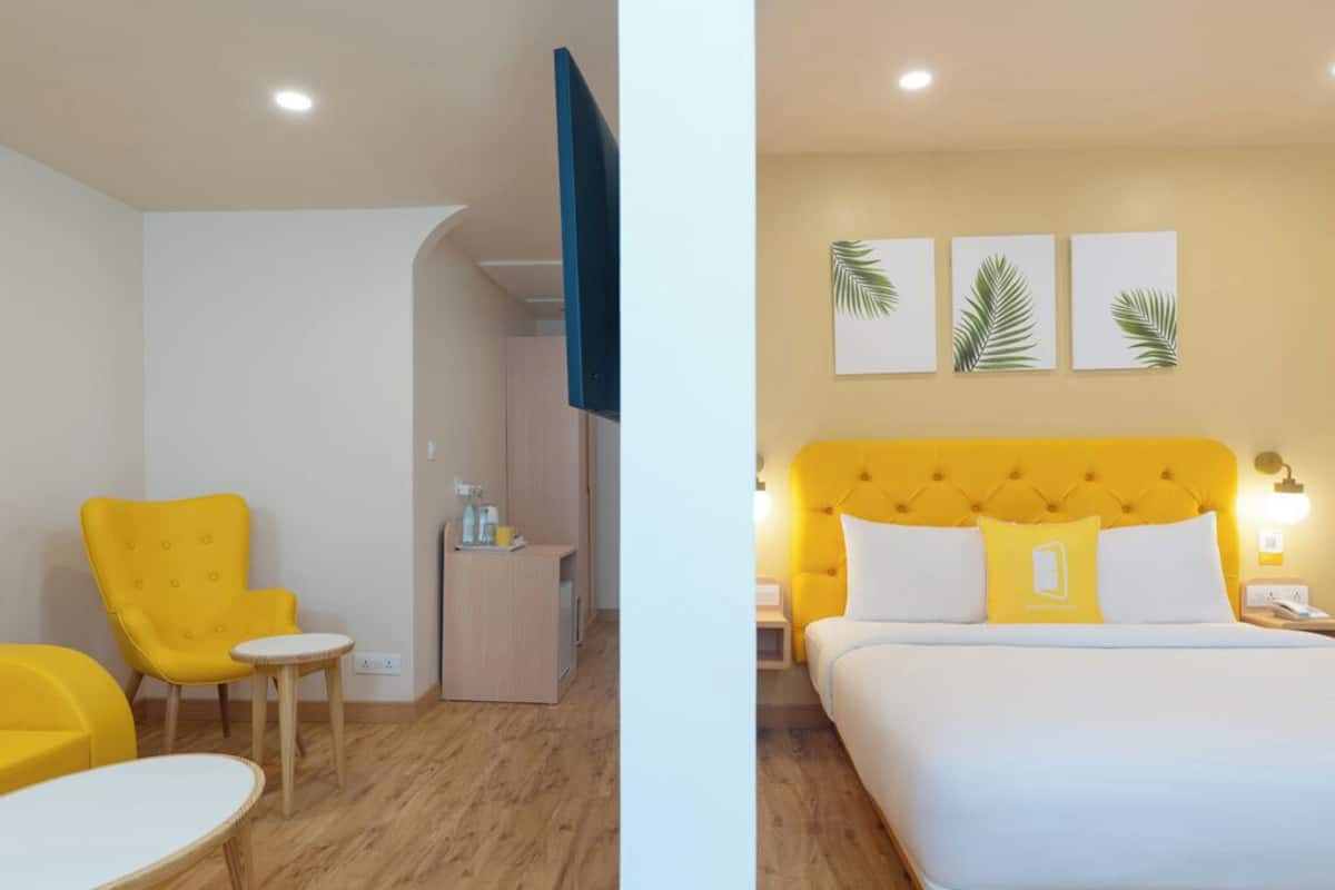 A room with a yellow armchair, a small round table, a bed with a yellow headboard, and a nightstand with a lamp.