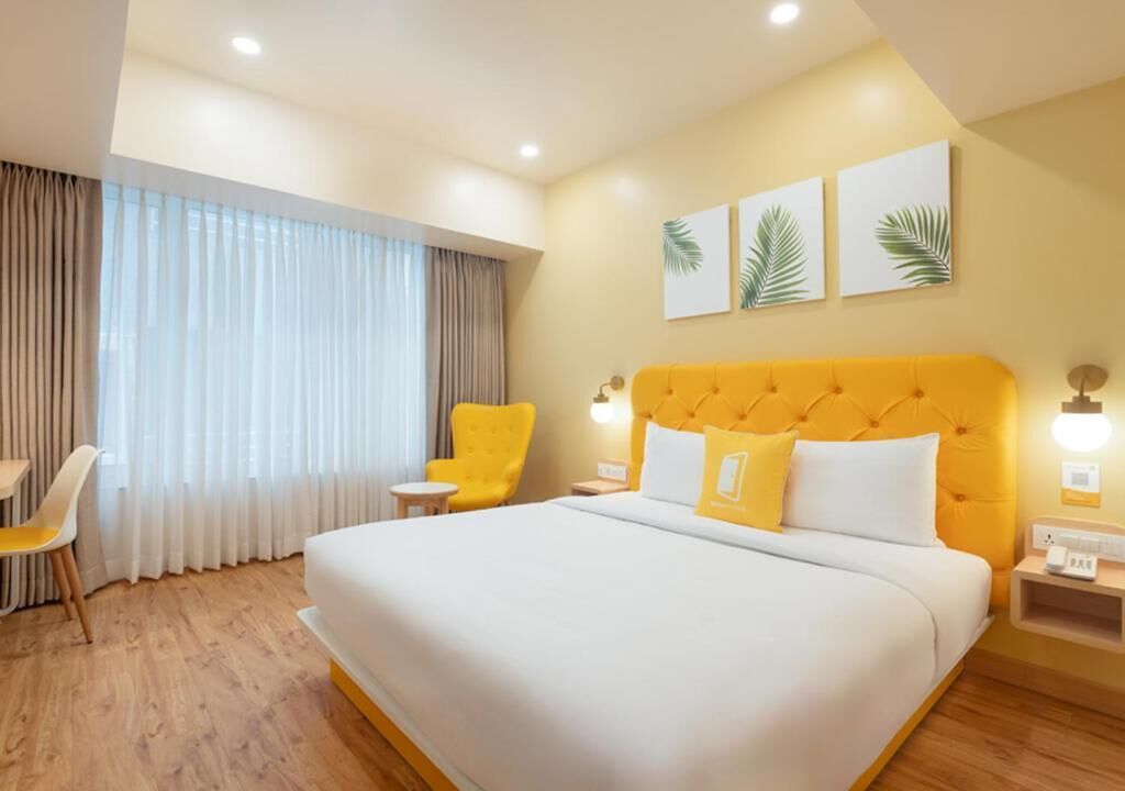 Une chambre d’hôtel avec un grand lit, un fauteuil jaune, une petite table et une fenêtre avec des rideaux.