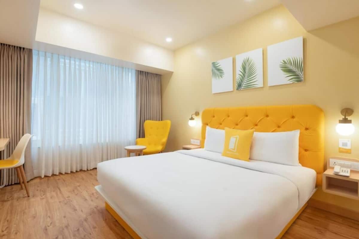 A hotel room with a large bed, a yellow armchair, a small table, and a window with curtains.