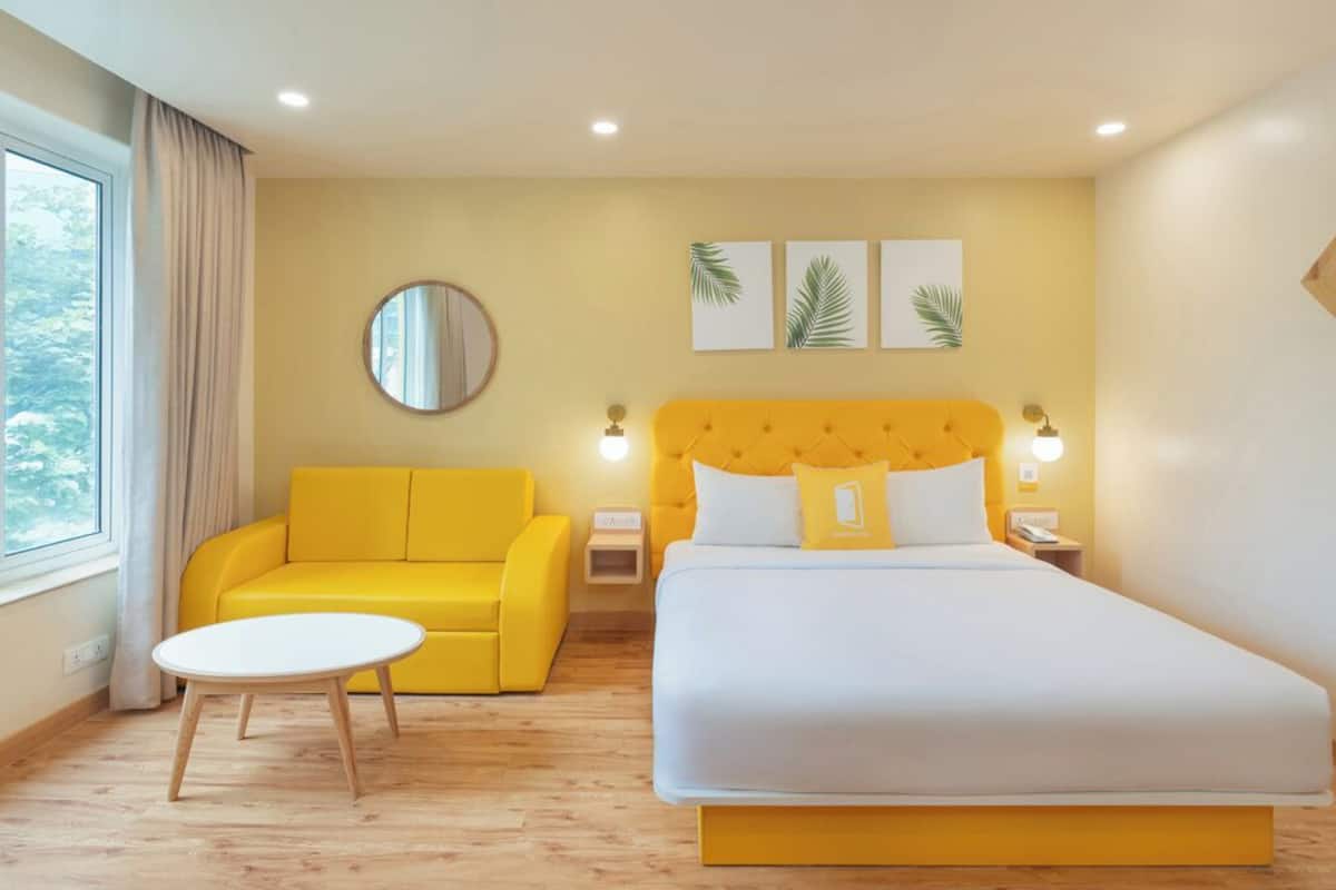 A hotel room with a yellow sofa, a round mirror, a bed with white bedding, and three wall-mounted light fixtures.