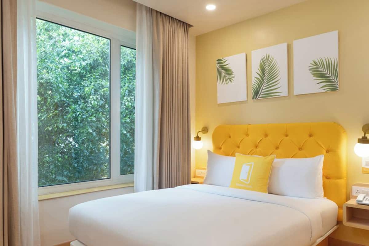 A hotel room with a bed, a window with curtains, a yellow headboard, and three palm leaf wall art pieces.