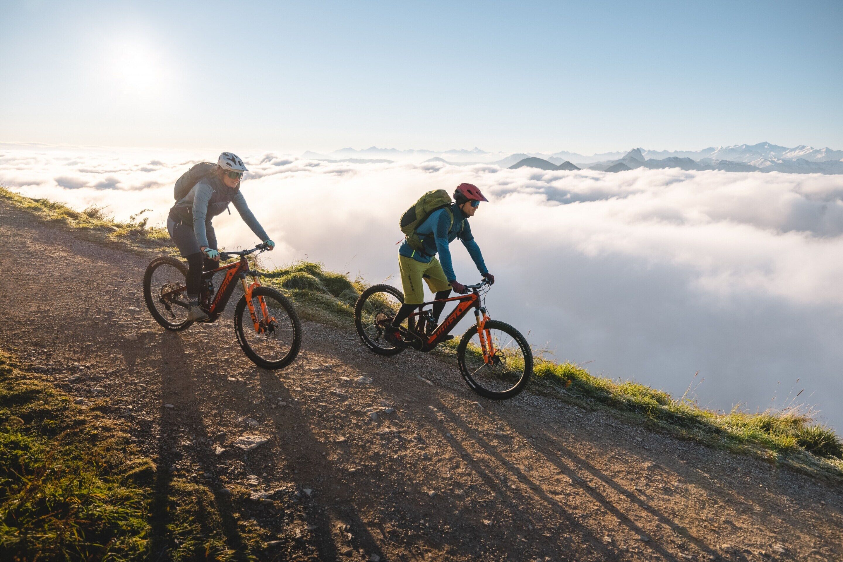 Deux cyclistes sur un sentier de montagne, avec une vue sur des sommets enneigés et des nuages en contrebas.