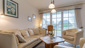 Ein Wohnzimmer mit einem beigen Sofa, einem Holztisch und einem Sessel. An der Wand hängt ein Gemälde, ein Fenster mit Vorhängen und Blick auf Häuser.