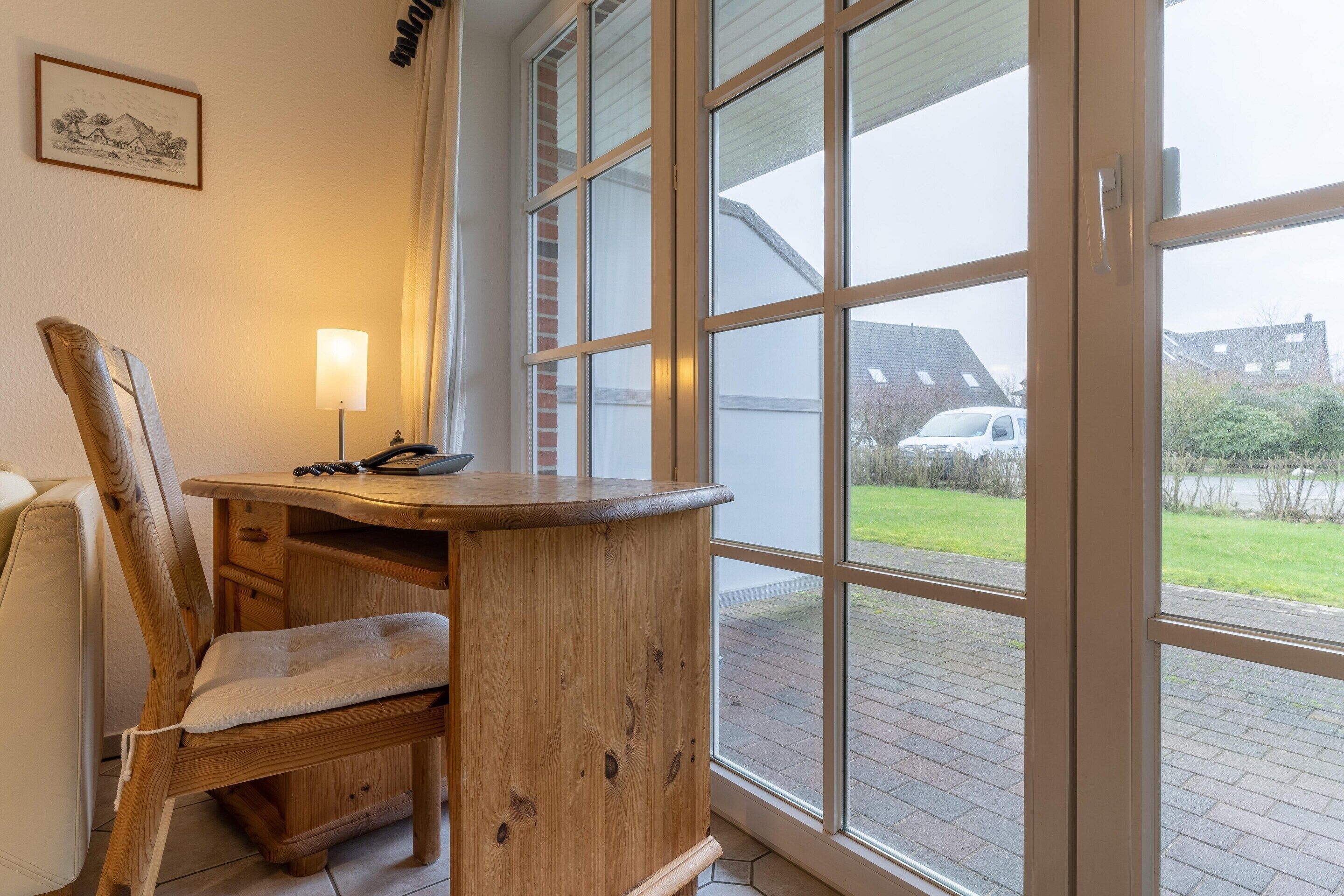 A room with a wooden desk and chair, a lamp, and a large window with a view of a brick pathway and buildings.