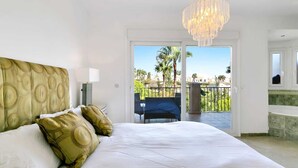 A bedroom with a large bed, a chandelier, and a balcony with a view of palm trees.
