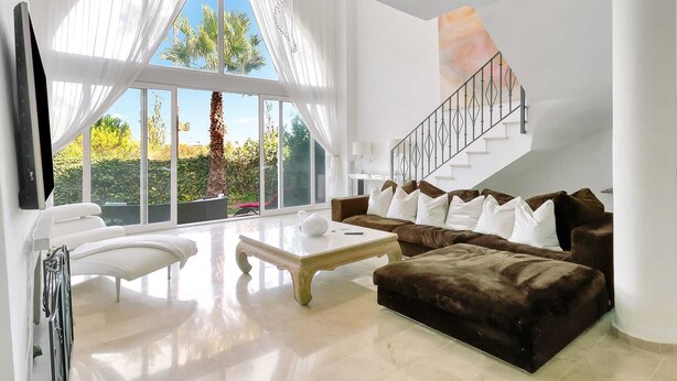 A spacious living room with a large window, a brown sofa, a white coffee table, and a staircase.