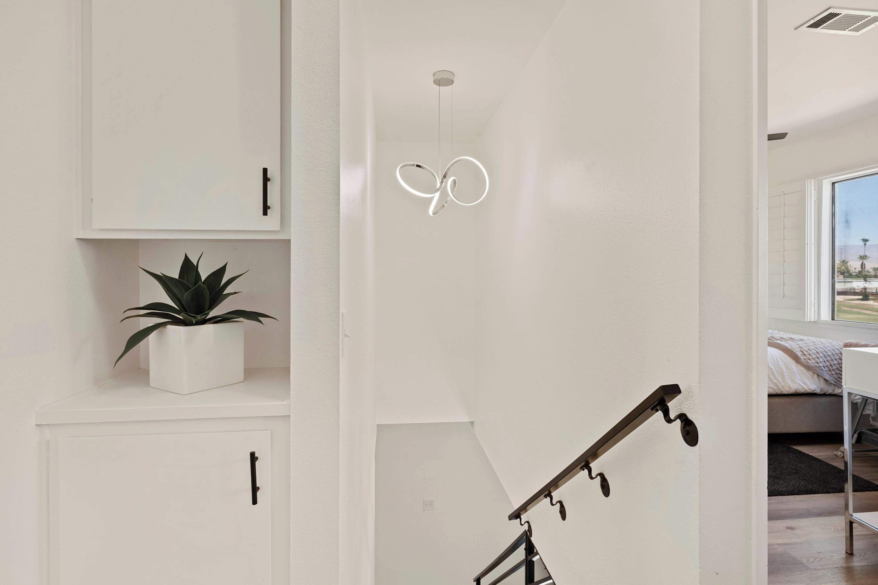 A modern interior with white cabinetry, a potted plant, a unique light fixture, and a staircase.