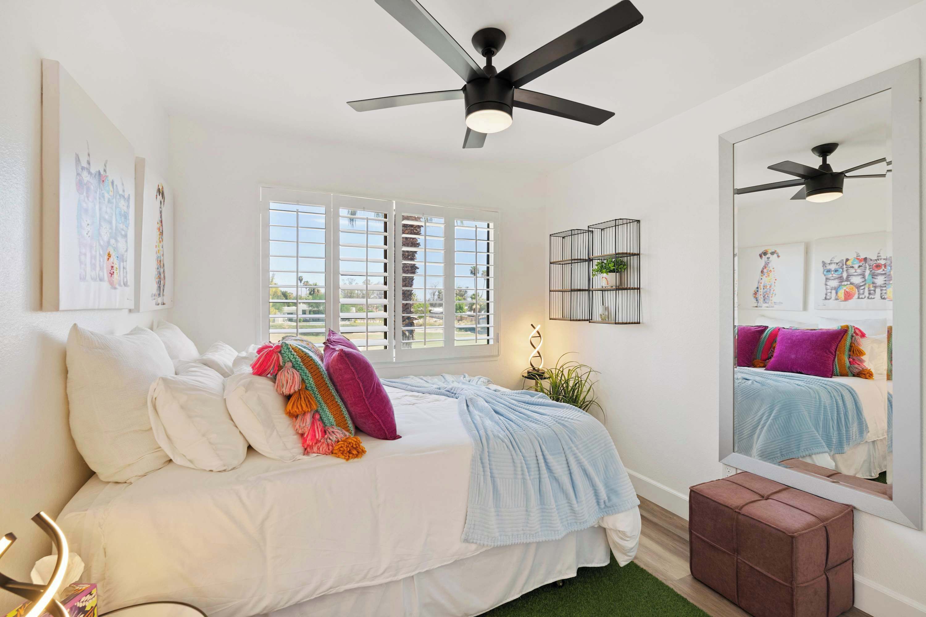 A bedroom with a bed, a ceiling fan, a window with blinds, a bedside table with a plant, and a mirror reflecting the bed.