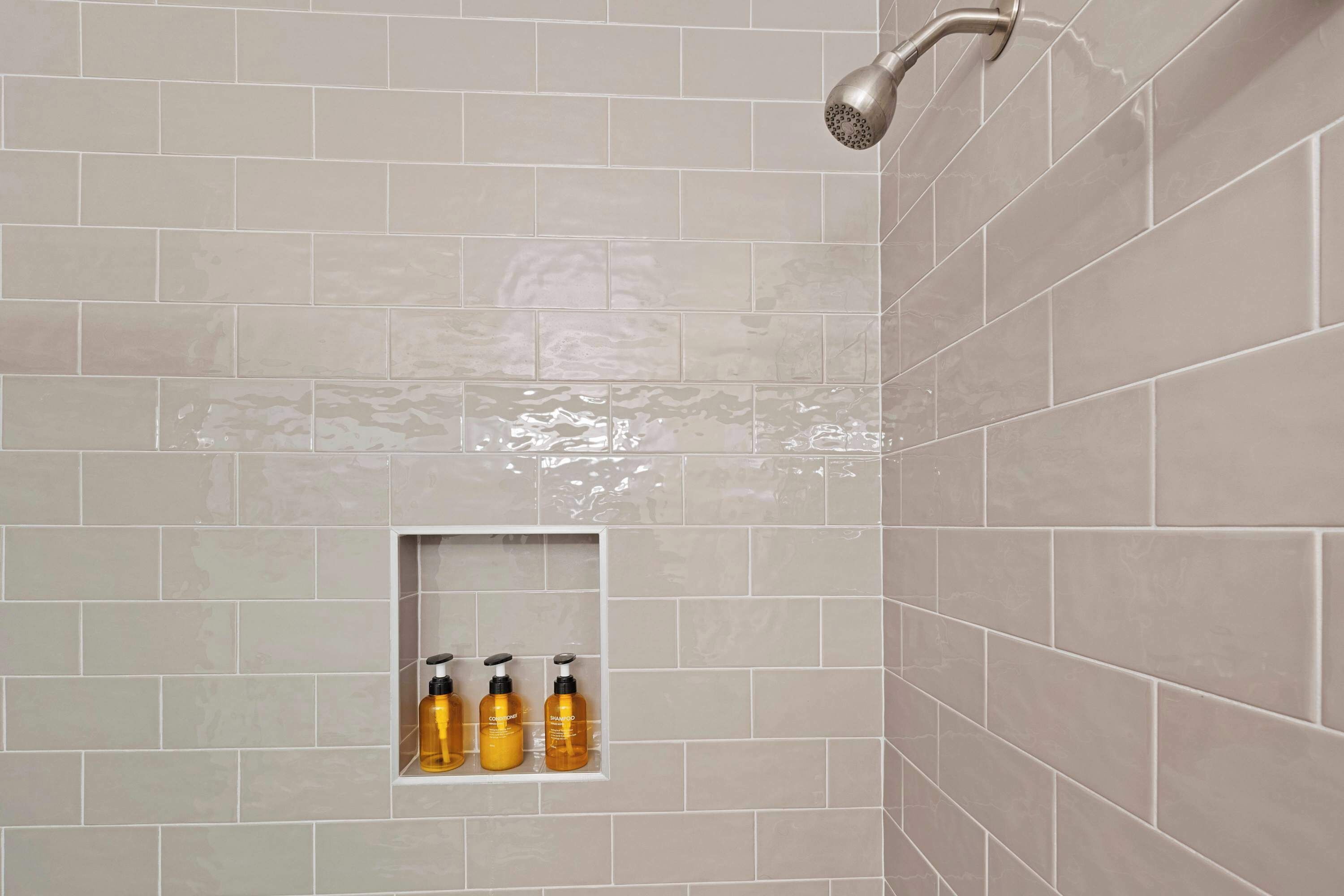 A shower area with beige tiles, a showerhead, and a shelf with three bottles.