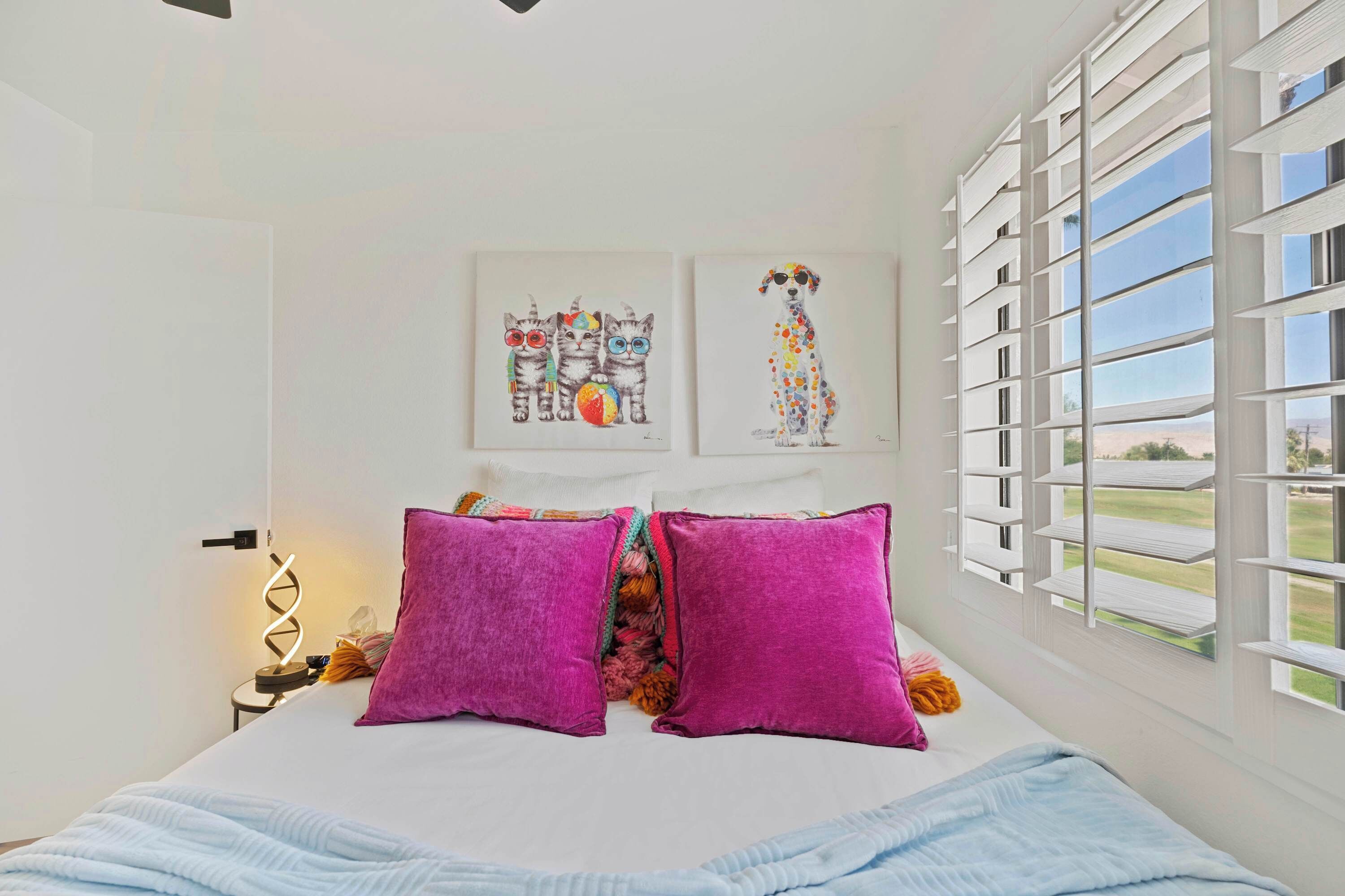 Une chambre à coucher avec un lit, deux oreillers, un tableau décoratif au mur, une lampe et une fenêtre avec des stores.