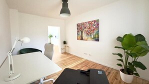 Ein Zimmer mit weißem Schreibtisch, einer schwarzen Massageliege, einem Blumentopf und einem farbenfrohen Gemälde an der Wand.