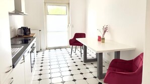 Eine kleine, moderne Küche mit weißer Arbeitsplatte, einem Herd und einem Spülbecken. Zwei rote Stühle und ein weißer Tisch mit einer Blumenvase stehen dort. Der Boden ist mit einem schwarz-weißen sechseckigen Fliesenmuster ausgelegt.