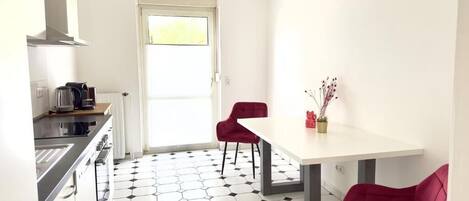 Een kleine, moderne keuken met een wit aanrecht, een fornuis en een spoelbak. Er staan twee rode stoelen en een witte tafel met een vaas bloemen. De vloer heeft een zwart-wit mozaïekpatroon van hexagonen.