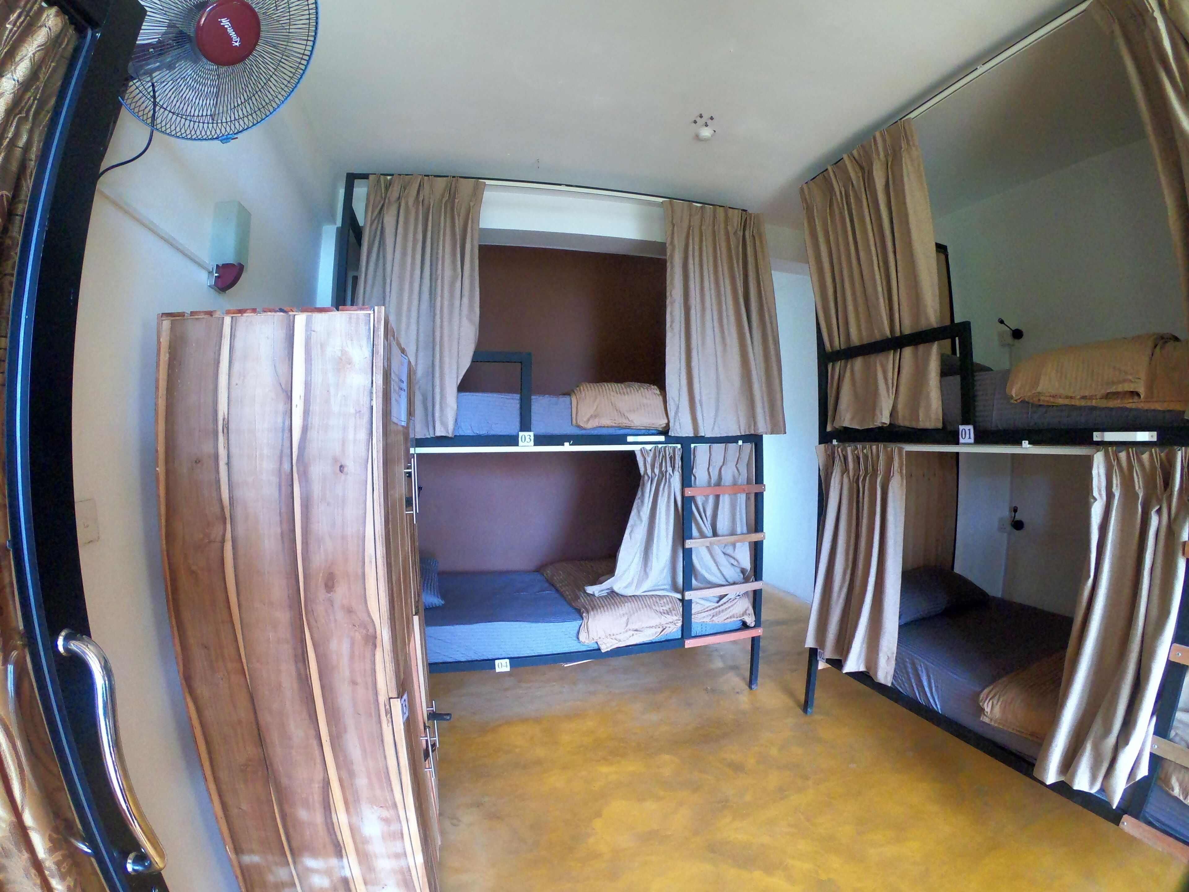 Une petite pièce avec des lits superposés, une armoire en bois, un ventilateur et des rideaux.