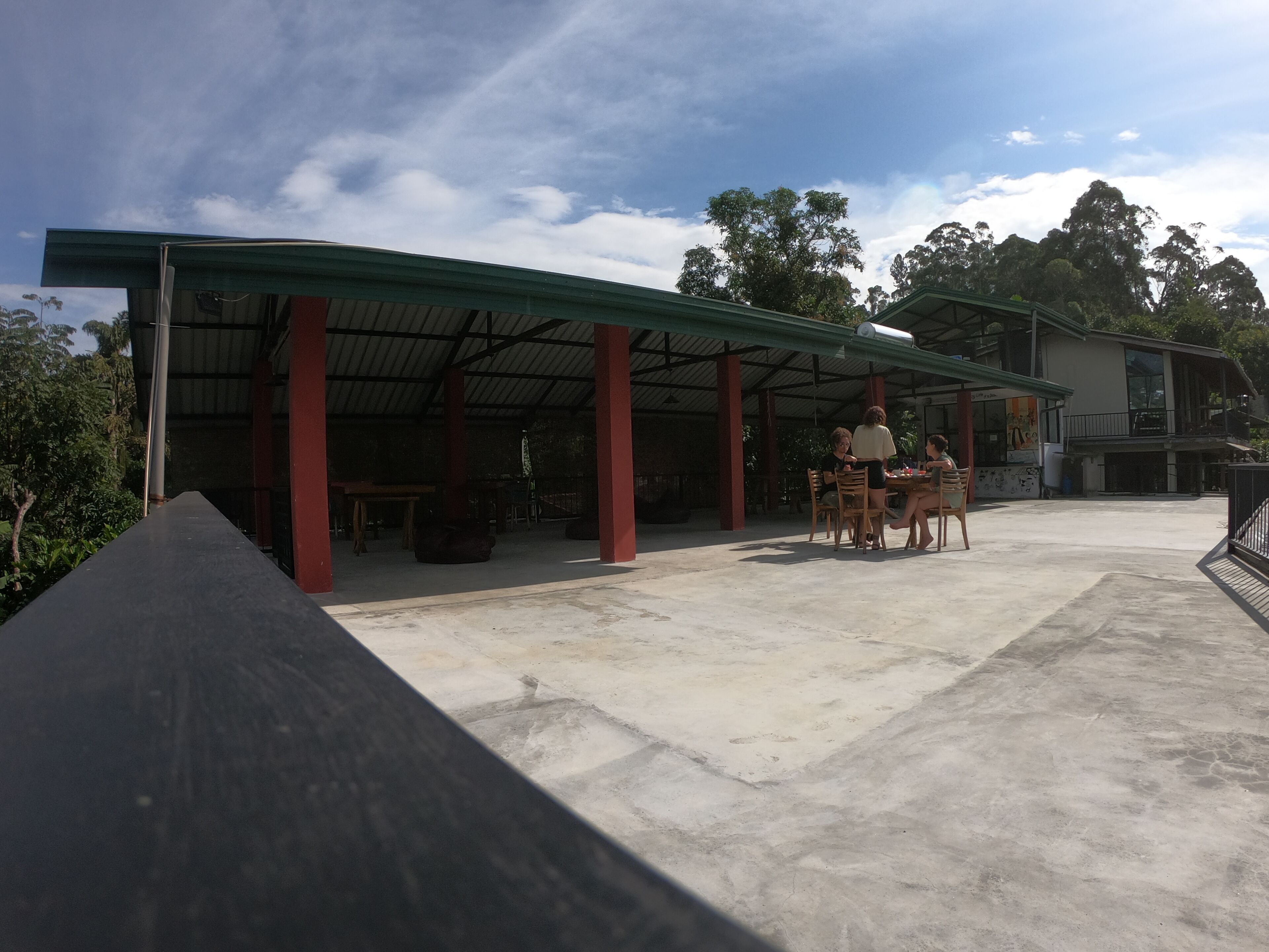 An outdoor covered area with a concrete floor, red pillars, and a table with chairs.